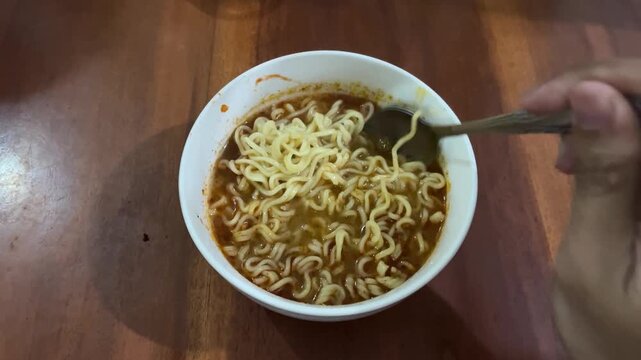 Hand using a spoon to stir hot spicy instant noodles in a white bowl on a wooden table, quick meal preparation footage.