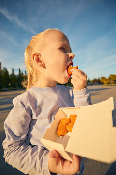 Hungry young girl biting nugget from open takeaway box in sunny park. Food enjoyment, childhood leisure concept.