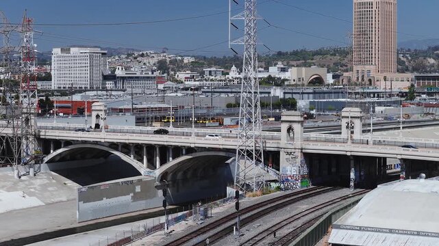 Aerial view tracks the historic First Street Bridge spanning across the concrete Los Angeles River channel with railway tracks, transmission towers, and downtown transit infrastructure.