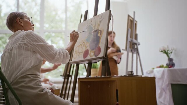 Senior Caucasian woman painting still life canvas on easel during art class. Ideal for promoting active retirement, adult education, and creative leisure campaigns