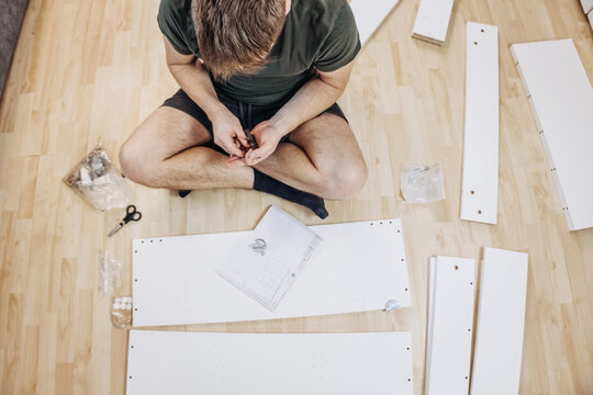 Man sitting on wooden floor reading instruction manual while assembling white wooden furniture in modern living room top view DIY project