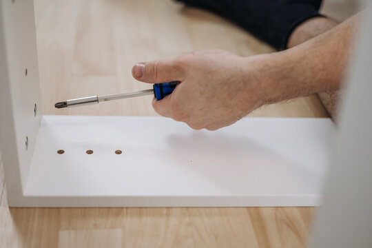 Hand holding blue screwdriver screwing bolt inside white furniture frame DIY assembly close up