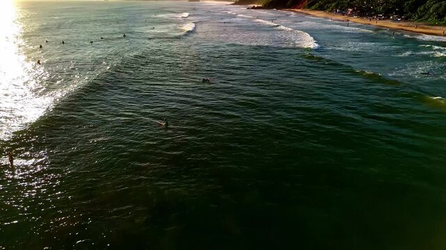 Aerial view of Madiha Beach with surfers in the ocean and waves rolling in under golden sunlight in Matara, Southern Province, Sri Lanka.