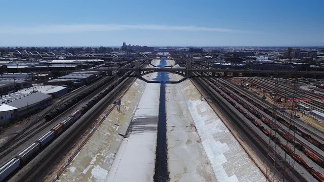 Aerial perspective looking along the concrete Los Angeles River flood control channel flanked by busy railway tracks, train yards, and historic bridges under clear daylight.