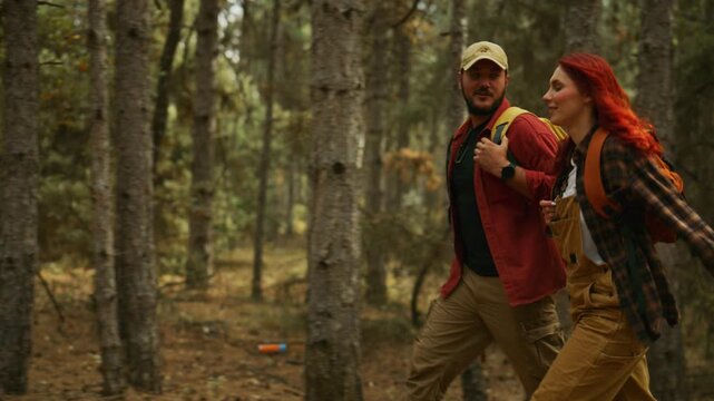 Couple walks side by side with backpacks through a sunny pine forest, talking and laughing during an autumn outdoor adventure.