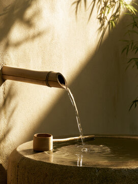 Bamboo water ladle garden fountain pouring water into stone basin under soft morning sunlight, peaceful landscaping scene with vertical space and natural shadow