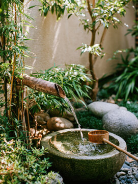 Bamboo water ladle garden fountain pours into stone basin, creating peaceful Japanese garden scene with greenery, river pebble, warm sunlight, and tranquil landscaping