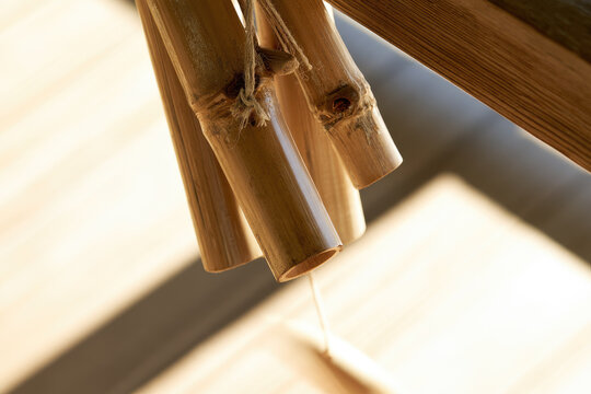 Bamboo wind chime tubes suspended from wooden porch overhang in warm morning sunlight, featuring natural hemp cord, hollow cane, weathered knots, soft shadow, and peaceful outdoor sound decor