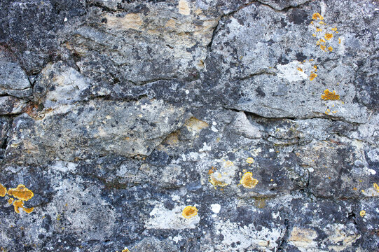 Old weathered stone wall covered with yellow and orange lichen, featuring rough surfaces, natural cracks and irregular rock patterns.