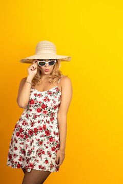 Woman pulling down sunglasses, making a kissy face while wearing a straw hat and floral dress on a yellow background