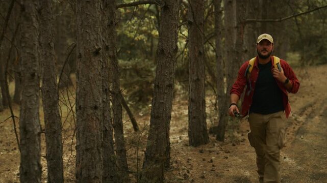Hiker walks along a dirt path through pine forest with a backpack and compass, wiping sweat from his forehead during a solo trek.