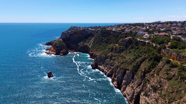 Turquoise Mediterranean waves breaking against rugged limestone cliffs and a sea stack, with hillside homes overlooking the coast of Costa Blanca, Spain
