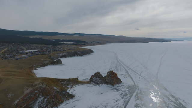 Seen from above, Shamanka Rock stands beside Olkhon Island, where snow and frozen water edge Lake Baikal near Khuzhir, Irkutsk Oblast, Russia, beneath a subdued winter sky with hills