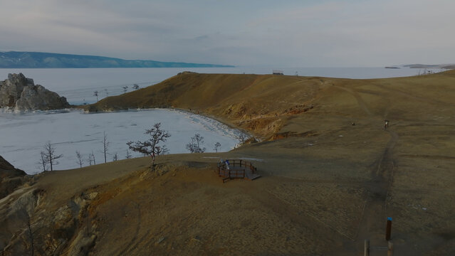 A high-angle winter photograph captures Shamanka Rock beside the frozen shore of Lake Baikal, with distant rugged mountains beneath a subdued sky on Olkhon Island, Irkutsk Oblast, Russia
