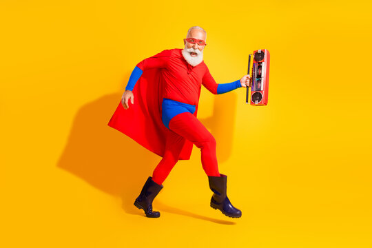 Energetic senior superhero dances with retro boombox against bright yellow background wearing red costume cape and mask