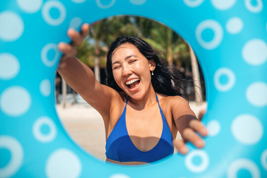 Happy young woman laughing through a blue inflatable ring on a tropical beach, summer vacation