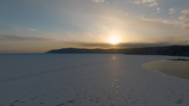 From above, winter sunset colors Lake Baikal near Listvyanka, where the Angara River flows out beside an icy shoreline and distant hills, under a calm evening sky in Irkutsk Oblast, Russia