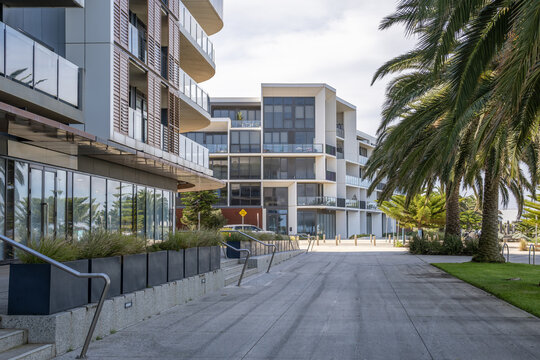 A paved pedestrian promenade alongside modern multi-story residential apartment buildings, featuring ground-floor glass shopfronts, outdoor steps with handrails. Modern mixed-use urban development
