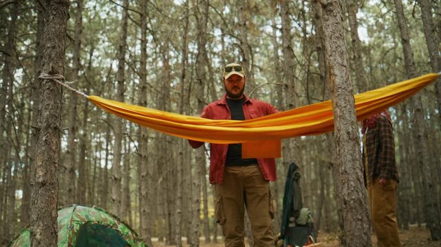 Camper and man adjust hammock fabric between pine trees at a forest campsite as the camera glides underneath during a camping trip.