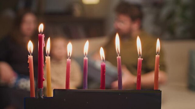 Lit Hanukkah menorah candles burning while blurred Jewish Caucasian family celebrating in background. Footage serves holiday advertising, cultural campaigns and religious storytelling projects