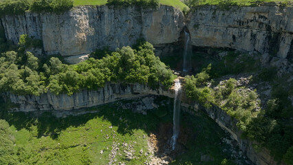 Fototapeta premium The Matlas waterfalls descend over a pale limestone ledge into a wooded ravine, where clear water winds among boulders and summer greenery beneath wide skies near Matlas, Dagestan, Russia