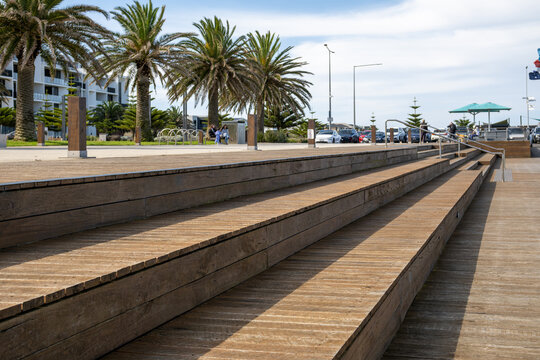 Wooden boardwalk decking and tiered seating steps at Wyndham Harbour in Werribee South, Melbourne, Australia,&nbsp;with tall palm trees, public amenities, and modern residential apartments in background