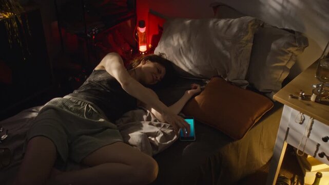 Caucasian young adult woman sleeping in bed, turning off ringing smartphone incoming call and going back to sleep. Perfect for digital detox and sleep deprivation campaigns