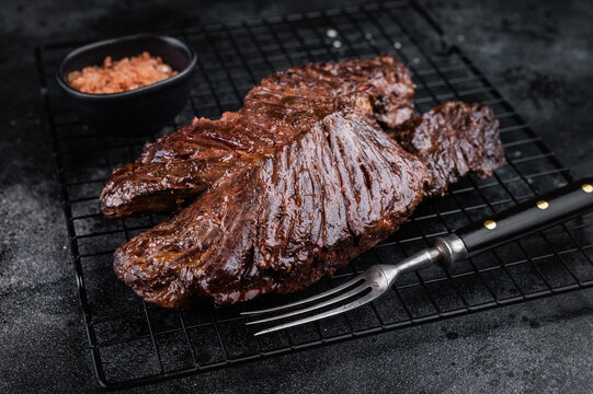 Grilled beef hanger steak onglet on metal rack with fork and coarse salt, dark stone background with copy space for barbecue concept.