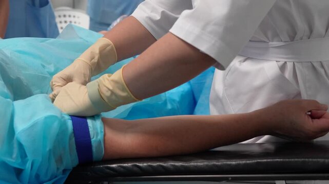 Medical nurse in sterile gloves fastening a tourniquet on a patient's arm to prepare for an intravenous catheter insertion or blood draw, a standard procedure inside a hospital room