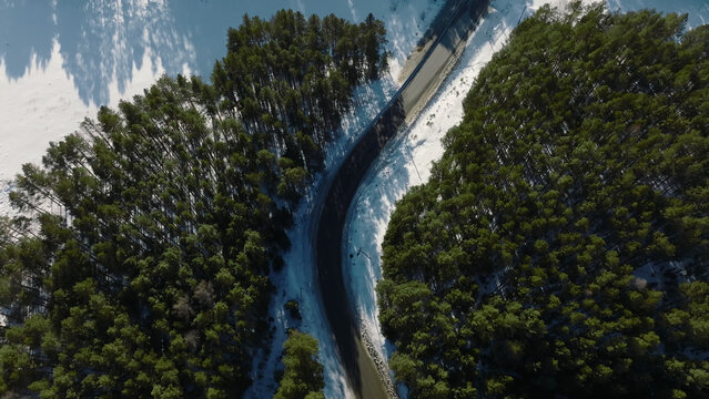 An elevated view follows a paved route edged with snow through dense evergreen woodland in the Ural Mountains, Russia, revealing a quiet winter passage across a wide remote landscape