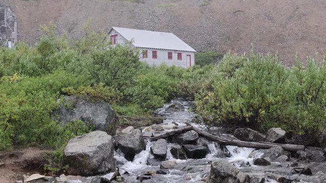 View of the historic Independence Mine visitor center nestled among lush green bushes and a rocky mountain stream in Palmer, Alaska, United States.