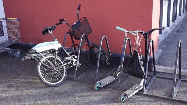Metal wave-form bike rack anchored beside red-walled building holds bicycle and kick scooter