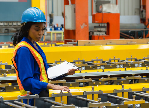 African Female Engineer Auditing Automated Metal Roll Forming Line : Professional quality inspector hardhat safety vest holding clipboard while check steel manufacturing machinery industrial plant