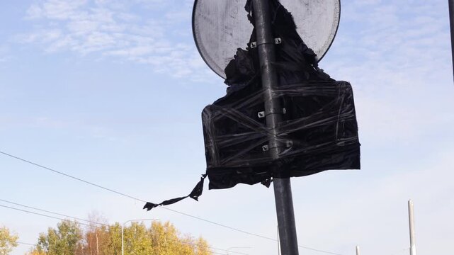 Round road sign partially wrapped in black protective covering mounted on metal pole amid urban street scene with autumn trees and utility wires
