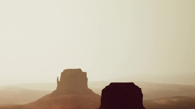The sun sets behind large rock formations in a desert. The sky has a light color as shadows stretch across the ground. The scene captures the natural beauty of the landscape.