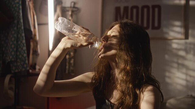 Young adult Caucasian woman drinking bottled water and pressing bottle to forehead feeling exhausted. Footage suitable for dehydration, summer heatwave awareness, and wellness video campaigns