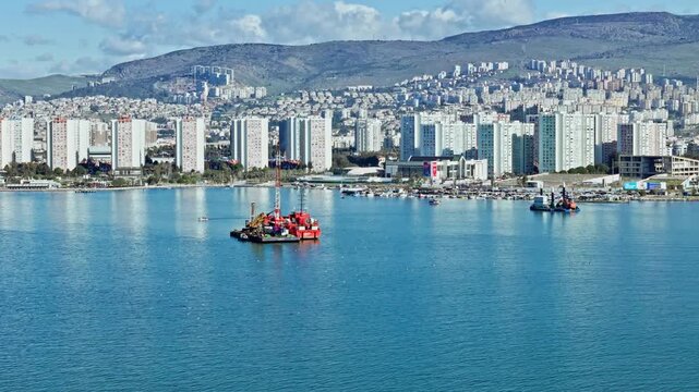 Floating crane barge working with dredging vessel near city waterfront and marina area. Aerial footage of coastal sediment removal and marine infrastructure maintenance
