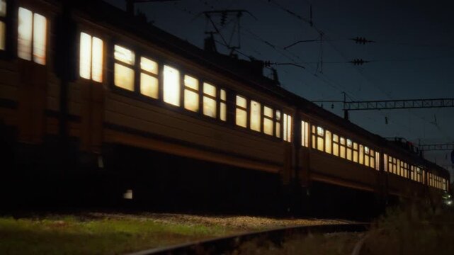 Night passenger rail transport - an electric train travels along electrified tracks with silhouettes of passengers visible through lit carriage windows and electric poles alongside