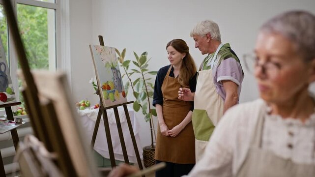 Senior Caucasian man painting still life on easel and discussing artwork with female teacher in art class. Footage ideal for promoting active aging and adult education programs