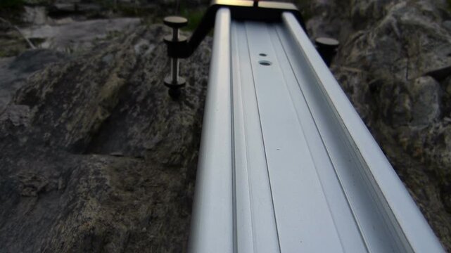 Close up camera tracking shot along an aluminum video slider over river rocks. Detailed backstage view of professional film equipment setup outdoors under bright sunshine during a media production.