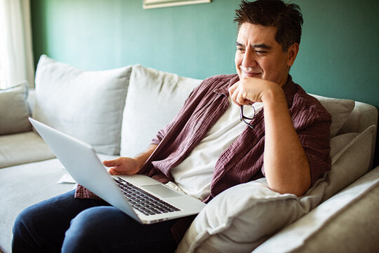 Smiling man holding glasses using laptop on sofa at home