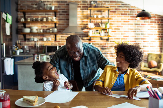 Man laughing with two children making paper airplanes at kitchen table