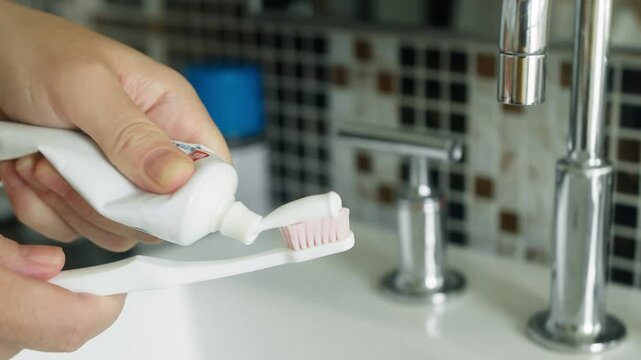 Close-up of a person squeezing toothpaste from a tube onto the bristles of a toothbrush beside a bathroom sink. Clean modern setting with chrome faucet and tiled background.