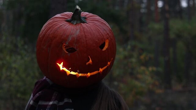 Spooky Halloween atmosphere with pumpkin-headed character in twilight forest. Bright orange Jack-lantern is located instead of the girl's head, it glows from the inside. Woman in Halloween costume