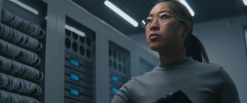 Young Female IT Specialist Reviews Technical Data on Tablet in Modern Server Room. Concept of Digital Infrastructure Monitoring, System Diagnostics, and Troubleshooting Potential Network Issues.