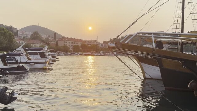 Static shot of a marina full of yachts near Dubrovnik, Croatia, with a bright orange sun setting at the center of the frame and boats gently rocking on the water. Original unedited 4K clip.