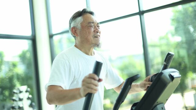 Elderly asian man exercising on a piece of gym equipment experiences sudden chest pain heart attack clutching his chest in agony due running overexertion.
