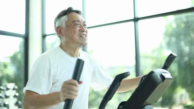 Elderly asian man exercising on a piece of gym equipment experiences sudden chest pain heart attack clutching his chest in agony due running overexertion.