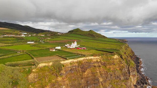 Ferraria Lighthouse stands among green coastal fields on Sao Miguel, Azores, Portugal, near Atlantic Ocean. White tower with red lantern anchors rural island travel scenery