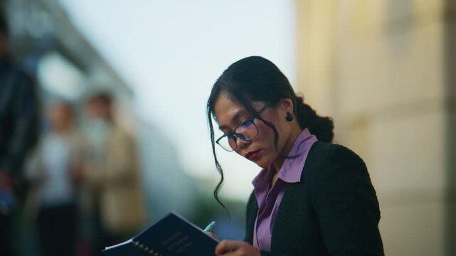 An Asian business woman is focused on planning and analyzing documents outdoors. She is intently reviewing papers and taking notes in a professional urban setting.